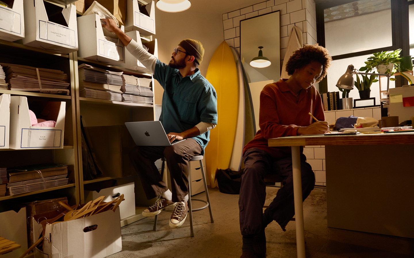 Osoba sedící za stolem píše tužkou do zápisníku, jiná osoba sedí vedle poličky s MacBookem na klíně a pravou rukou sahá pro složku.