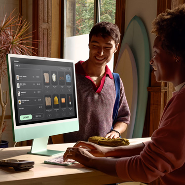 Zaměstnanec sedící za přepážkou v prodejně se surfovými prkny a podobným zbožím dokončuje na iMacu nákup pro zákazníka, který stojí před přepážkou.