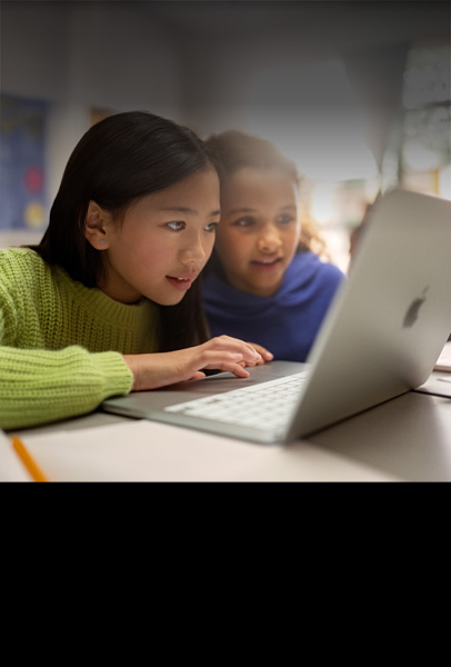 Two young students seated at a desk looking intently at MacBook as they collaborate in a classroom