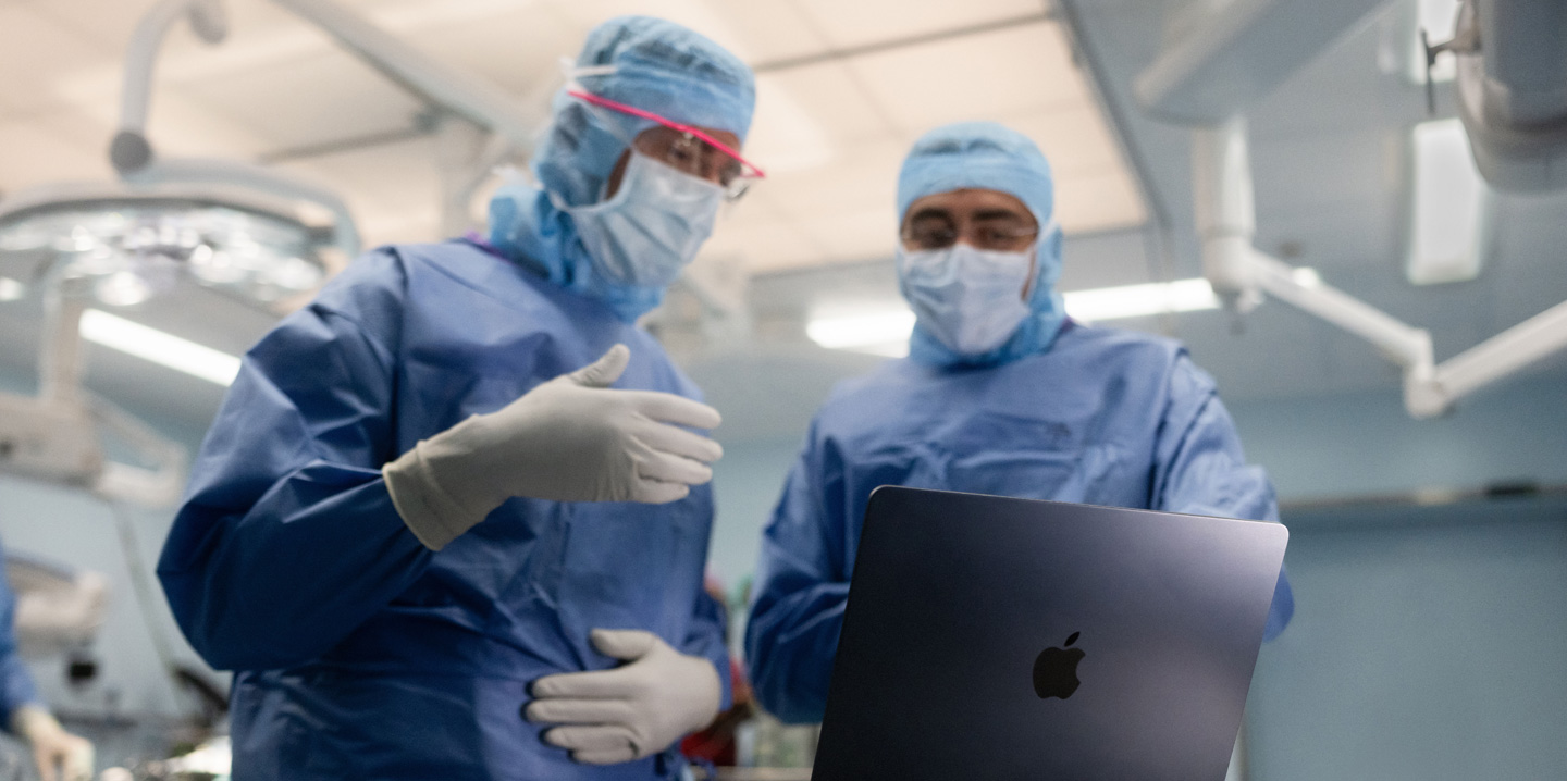 Deux chirurgiens dans une salle d’opération regardent un MacBook, l’un d’eux fait un geste de la main tandis que l’autre l’observe