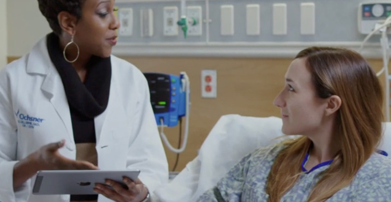 En læge med en iPad taler med en patient i en hospitalsseng.